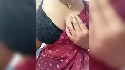 Media: Video of a woman's chest, showcasing a black lace bra under a red floral sari. Her left hand covers her right breast, revealing a gold pendant necklace. The background is blurred, focusing on the sari and bra.