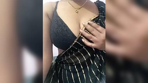 Media: Video of a woman with medium brown skin, wearing a black lace bra with gold trim, partially unbuttoned, revealing cleavage. She has gold jewelry and manicured nails. Background is blurred.