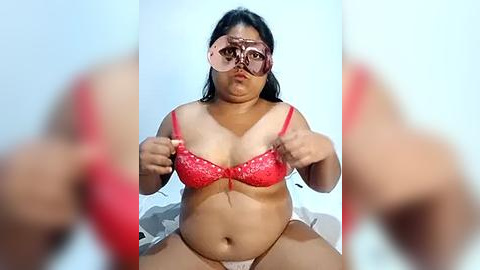 Media: Video of a plus-sized woman with medium brown skin, wearing a red lace bra and panties, with pink sunglasses, seated on a bed with a white sheet.