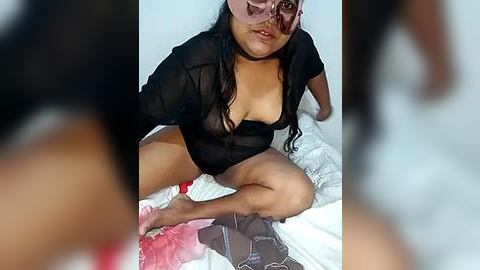 Media: A video of a plus-sized, dark-skinned woman with long hair, wearing a sheer black bodysuit, sitting on a bed with a white sheet, partially covered in red and white fabric. Her face is partially obscured by a white mask.