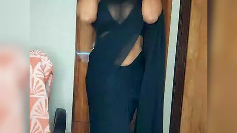 Media: Video of a person in a sheer black dress with a high slit, revealing a glimpse of their skin, standing in a room with wooden doors and a patterned towel.