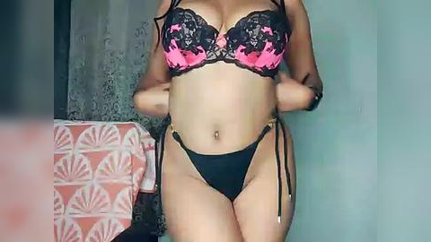 Media: Video of a light-skinned woman wearing a black lace bra with pink floral embroidery and matching black thong, standing in a dimly lit room with a patterned pink and white cloth on the left.