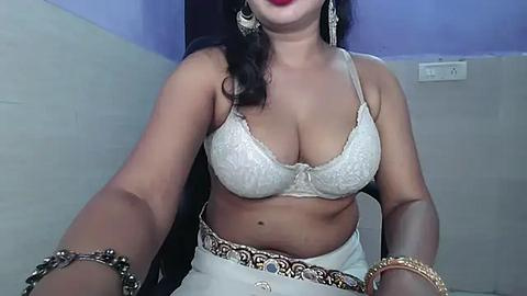 Media: Video of a curvaceous woman with medium brown skin, wearing a white lace bra and matching skirt, adorned with gold bling. She has long black hair and wears red lipstick.