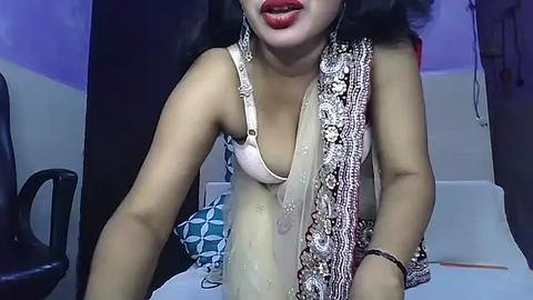 Media: Video of an Asian woman with medium skin tone, wearing a white bra and a detailed, gold-embroidered saree, sitting indoors with a dark chair and a blue-patterned cushion in the background.