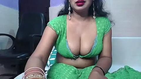 Media: Video of a curvy South Asian woman in a green, low-cut blouse and matching skirt, sitting on a bed. She has long, wavy black hair and wears silver earrings. The background includes a black chair and white walls.