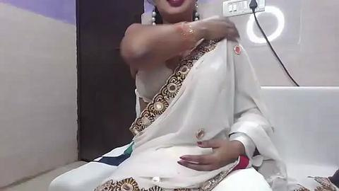 Media: Video of a woman in a white sari with intricate gold embroidery, seated in a modern bathroom with white tiles, taking a selfie, holding her arm.