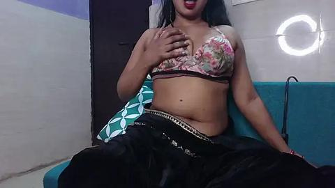 Media: Video of a young South Asian woman with medium brown skin, sitting on a teal couch. She wears a floral bra and black pants, with a green and white patterned pillow beside her.