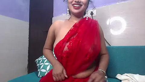 Media: Video of a smiling South Asian woman with medium skin tone and medium build, wearing a red saree, adorned with white floral earrings, seated on a teal sofa with green patterned cushions in a modern bathroom with tiled walls.