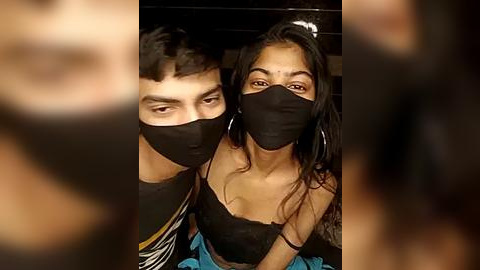 Media: Video of a young South Asian couple in a dimly lit room. Both wear black face masks, the man has short hair, and the woman has long, wavy hair. They are dressed casually; the man in a t-shirt, the woman in a black top.