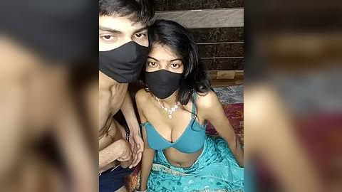 Media: Video of a young South Asian couple in a cozy, dimly lit room. The man, shirtless, has a mask and is behind the woman, who wears a turquoise bra and blue skirt.