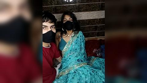 Media: Video of a young woman in a turquoise saree, wearing a black mask, sitting with a young man in a red shirt, both indoors.