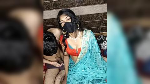 Media: Video of an Indian woman in a blue saree with red blouse, black face mask, and gold earrings, kissing a shirtless man with short black hair, in a rustic, tiled room.