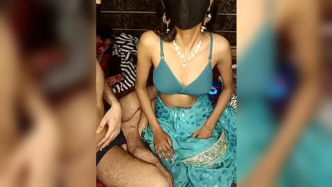 Media: A video shows an Indian woman in a teal saree, seated, with a black face mask, and a man behind her, partially visible, in a traditional setting.