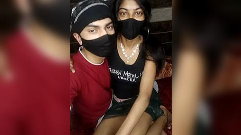 Media: Video of a young South Asian couple with medium skin tones, wearing black face masks, casual clothing, and sitting close on a red couch.