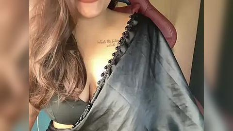 Media: Video of a woman with long, wavy brown hair, wearing a black lace bra with a silver chain, holding a black satin robe.