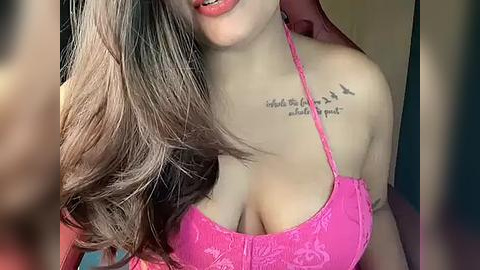 Media: Video of a young woman with long, wavy brown hair, wearing a pink lace bra, showcasing a tattoo on her shoulder. Her skin is light, and she has full lips.