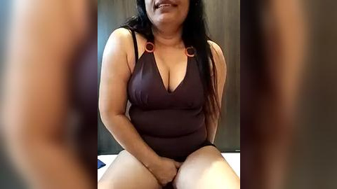 Media: Video of a smiling, plus-sized woman with long black hair, wearing a revealing, dark brown, halterneck swimsuit with orange circular decorations, sitting on a bed against a wooden backdrop.