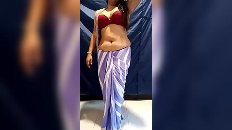 Media: Video of a South Asian woman in a red bra and white and purple saree, walking indoors, blurred figures in the foreground.