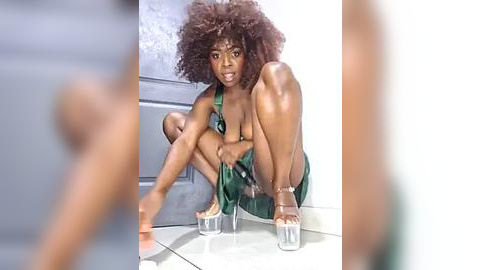 Media: Video of a Black woman with voluminous natural curls, wearing a green halter top and high-heeled platform shoes, crouched in a studio with white walls and reflective floor.