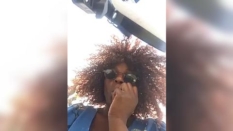 Media: Video of a Black woman with curly hair, wearing sunglasses, holding her face in her hands, in a blue shirt, looking out a car window.