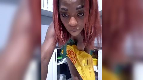 Media: Video of a black woman with shoulder-length, vibrant pink hair, leaning over a counter, wearing a green top. She holds a yellow container with orange spaghetti, in a modern kitchen.