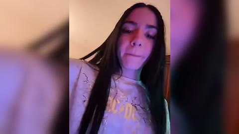 Media: Video of a young woman with long black hair, wearing a white shirt with a yellow design, standing in a dimly lit room with purple and pink lighting.