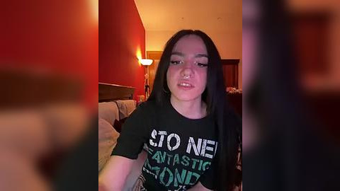 Media: Video of a young woman with long black hair, light skin, wearing a black T-shirt with \"STONED\" and \"ANASTIC\" in white and green letters. She is indoors, in a dimly lit room with red walls, a bed, and a lamp.