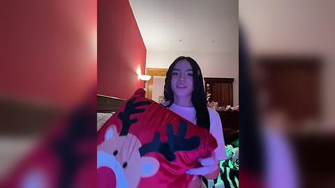 Media: Video of a smiling young woman with long black hair, wearing a white long-sleeve shirt, holding a red blanket with white and black cartoon characters. Background features a red wall, wooden furniture, and a dimly lit room.