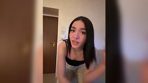 Media: Video of an Asian woman with straight black hair, wearing a black tank top, kneeling on a couch in a simple, beige-walled room.