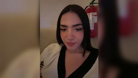 Media: Video of a young woman with medium skin tone and straight black hair, wearing a white robe with black accents, and a septum piercing. She's in a dimly lit room with a red fire extinguisher on the wall.