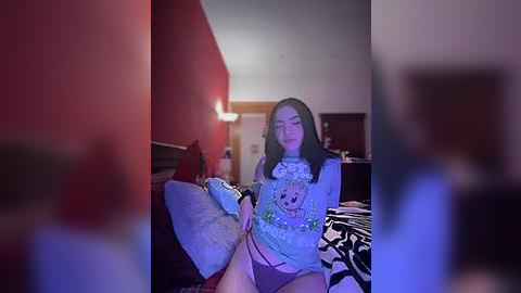 Media: A blurred video of a young woman in a colorful T-shirt and pink panties, sitting on a bed in a dimly lit room with red walls and white ceiling.