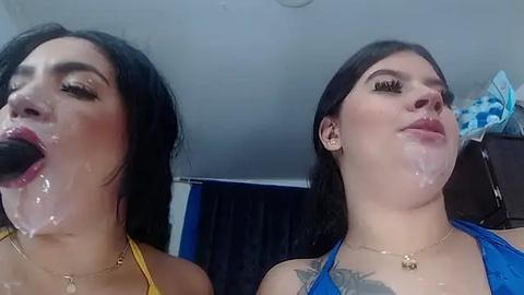 Media: Video of two young women with long black hair, wearing yellow and blue tank tops, with semen on their faces, standing in a dimly lit room.