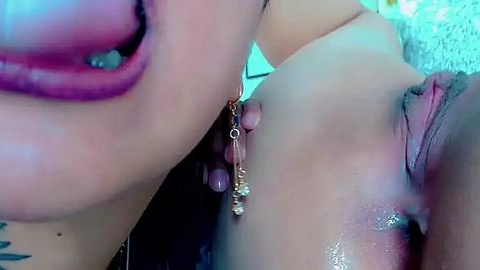 Media: Video of a close-up of a woman's face and chest, with a man's penis and testicles touching her breast. Her lips are painted pink, and she has a gold necklace. Background is blurred.