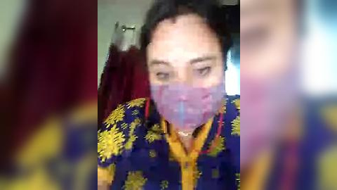 Media: Video of a man with a mask, wearing a blue shirt with yellow patterns, standing in a blurry room with red and blue clothes hanging.