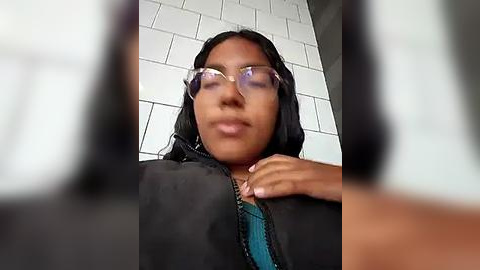 Media: Video of a woman with medium brown skin and long black hair, wearing glasses, a black jacket, and a teal top. Background features white tiled walls.