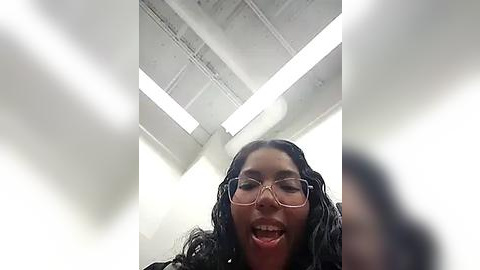 Media: Video of a young Black woman with curly hair, wearing glasses, laughing with her mouth open, in a brightly lit, white ceiling room with fluorescent lights.