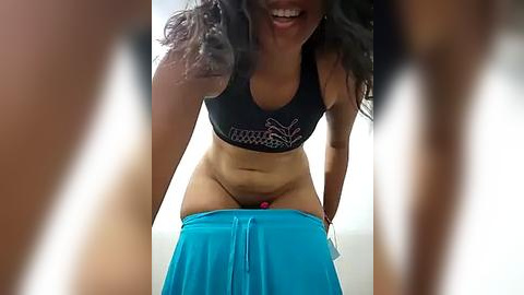 Media: Video of a smiling woman with medium-dark skin, long curly hair, wearing a black sports bra and blue yoga pants, bending over, captured from a low angle.