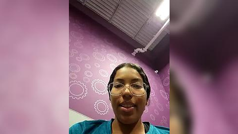 Media: Video of a smiling woman with medium brown skin, wearing glasses and a teal shirt, standing in a room with purple walls decorated with white gears and a ceiling with fluorescent lights.