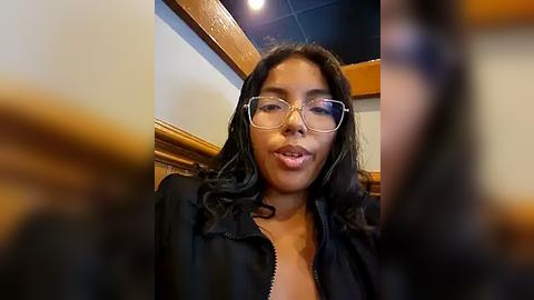 Media: Video of a Black woman with shoulder-length black hair, wearing clear-rimmed glasses and a black jacket, smiling in a dimly lit, wooden-paneled room.