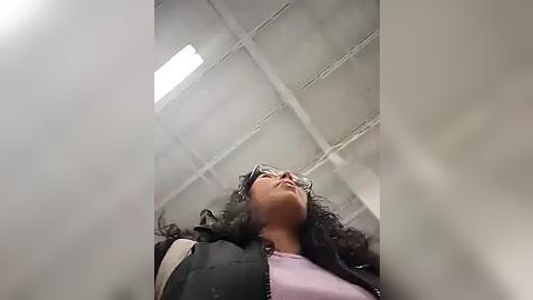 Media: Video of a young woman with curly black hair, wearing sunglasses, a black jacket, and a light pink shirt, looking upwards inside a dimly lit, grid-patterned ceiling.