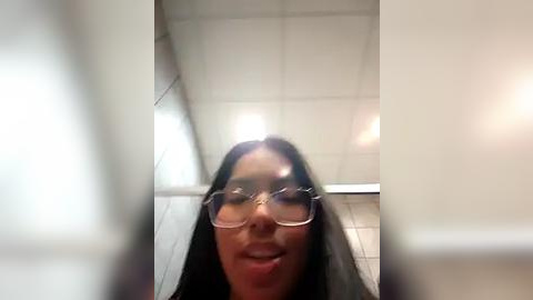 Media: Video of a woman with long dark hair and glasses, standing in a white-tiled bathroom with fluorescent lights. Her face is slightly blurred, creating a surreal, dreamlike effect.