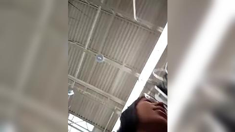 Media: Video of an industrial ceiling with corrugated metal panels and fluorescent lights, taken from a low angle, capturing a smiling person in the foreground with short black hair.