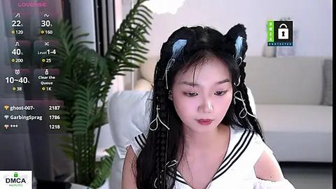 Media: Video of a young East Asian woman with long black hair, wearing a white sailor collar top and black cat ears, seated indoors. Background includes a modern living room with a couch, fern, and digital interface.