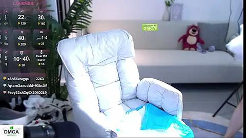 Media: A video of a modern living room with a white armchair, a gray sofa, and a plush red teddy bear. A virtual camera overlay shows live viewers' names and emojis.