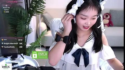 Media: Video of an Asian woman with long black hair in a maid outfit, holding a phone, in a modern living room with a potted plant.