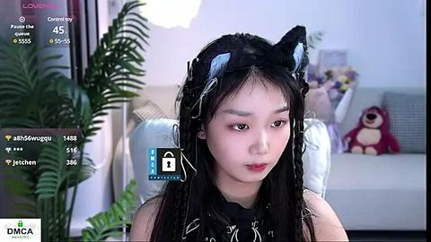 Media: Video of a young Asian woman with fair skin, wearing a black top, black cat ears, and black hair, seated in a modern living room with a plush toy and plants.