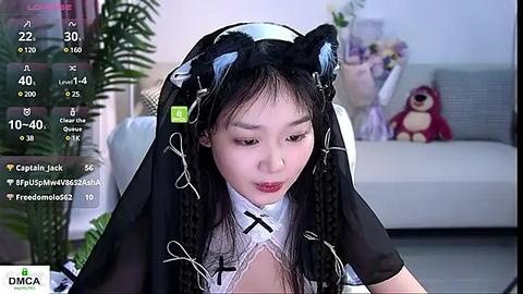 Media: Video of an Asian woman in a black cat ear headband, white maid outfit, and black gloves, sitting in a modern living room with a plush toy in the background.