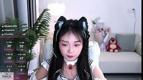 Media: Video of an East Asian woman in a white dress with black cat ears, seated indoors. Background features a light-colored couch with stuffed toys, a plant, and a wall light.