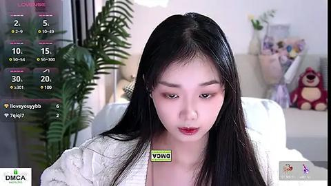 Media: Video of a young Asian woman with long black hair, fair skin, and red lipstick, wearing a white sweater. Background features a living room with a potted plant, stuffed toys, and a soft couch.