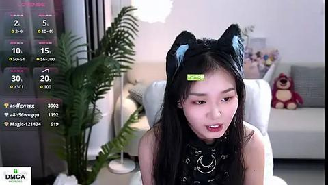 Media: Video of an Asian woman with long black hair, wearing a black cat ears headband, a black top, and a silver necklace. She's in a modern living room with a couch and ferns.